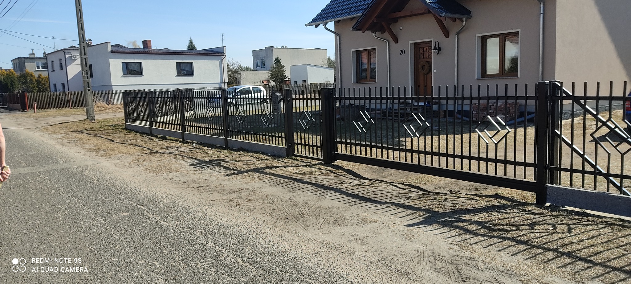Czarna, metalowa brama przesuwna i ogrodzenie z dekoracyjnymi elementami w kształcie rombów przed domem z numerem 20.