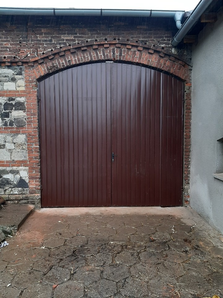 Dwuskrzydłowa, brązowa brama garażowa z pionowymi panelami, osadzona w ceglanym łuku, z widocznymi fragmentami starego muru i brukowanej nawierzchni przed bramą.
