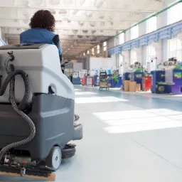 Operator czyszczący podłogę w hali produkcyjnej za pomocą automatycznej maszyny czyszczącej z widocznymi szczotkami i ssawką