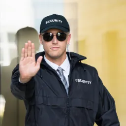 Mężczyzna w czarnej kurtce z napisem 'SECURITY' na tle przeszklonego wejścia, podnosi dłoń w geście stop.