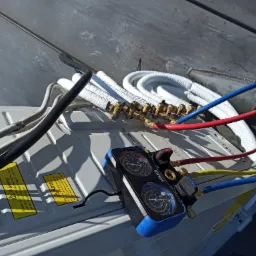 Podłączenie przewodów chłodniczych do klimatyzatora z użyciem manometru serwisowego; widoczne białe izolowane rury, kolorowe przewody i naklejki ostrzegawcze na obudowie urządzenia.