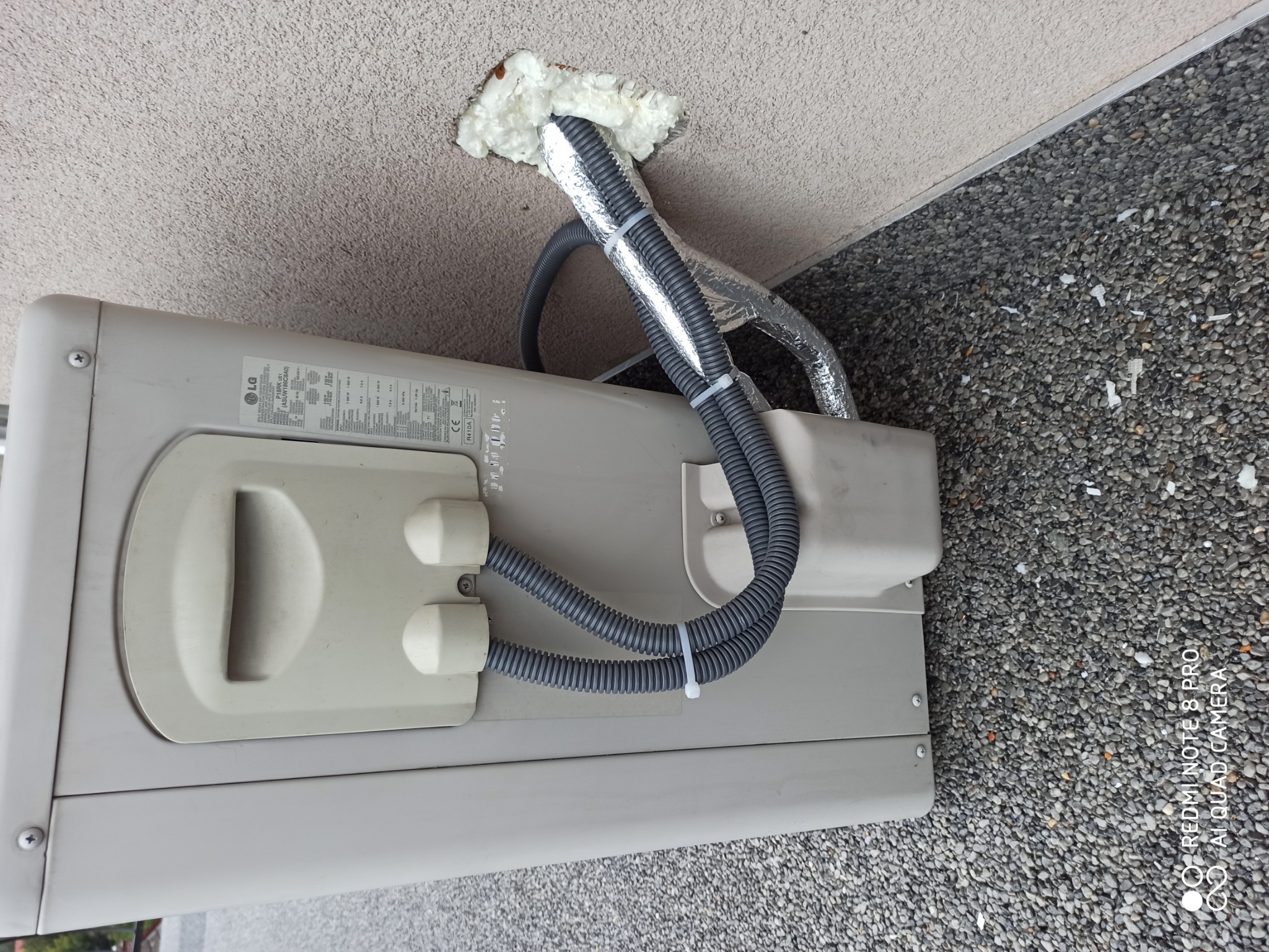 Zewnętrzna jednostka klimatyzacji LG zamontowana na ścianie budynku, z widocznymi przewodami w peszlach przymocowanych opaskami zaciskowymi i rurami owiniętymi folią aluminiową, wchodzącymi...