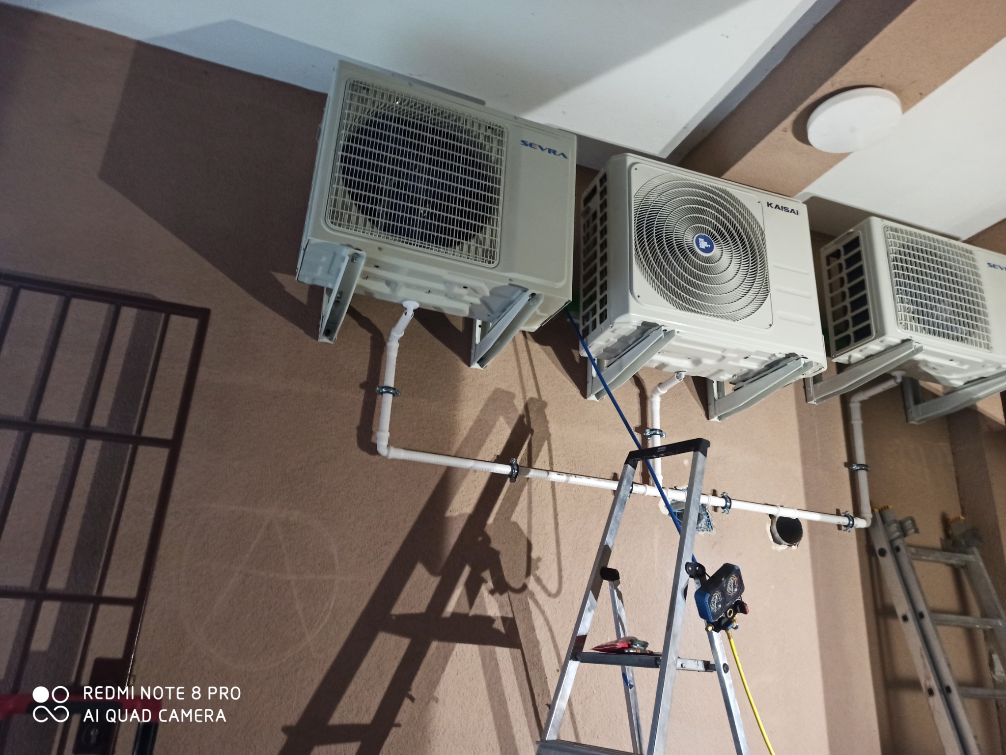 Trzy klimatyzatory marki Seyra i Kaisai zamontowane na ścianie, połączone rurami, widoczne drabiny i narzędzia serwisowe, w tle beżowa ściana.