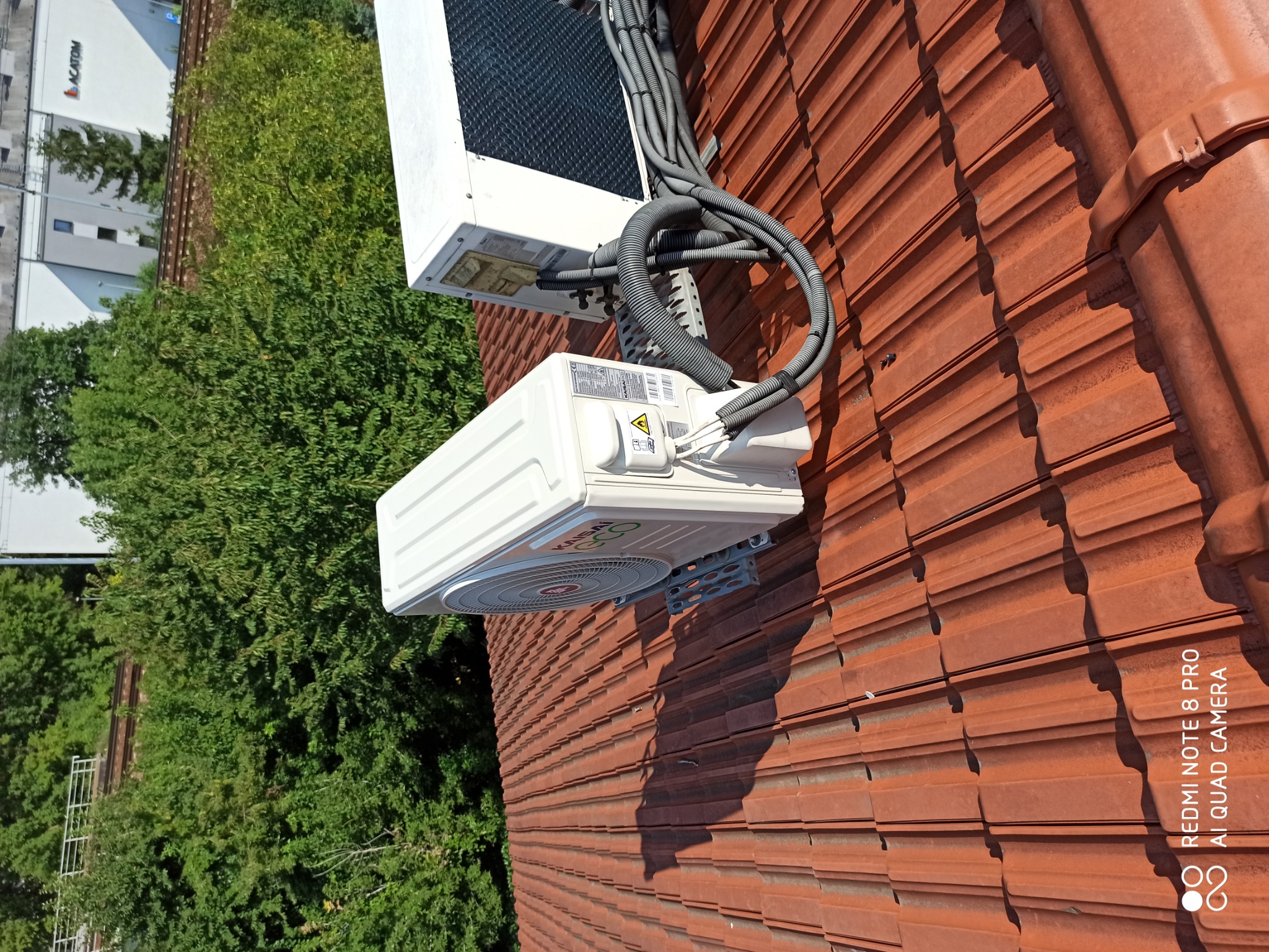 Zewnętrzna jednostka klimatyzacji marki Kaisai zamontowana na czerwonym dachu z dachówki ceramicznej, widoczne kable w peszlach prowadzące do jednostki zewnętrznej, w tle zieleń drzew i budynek.