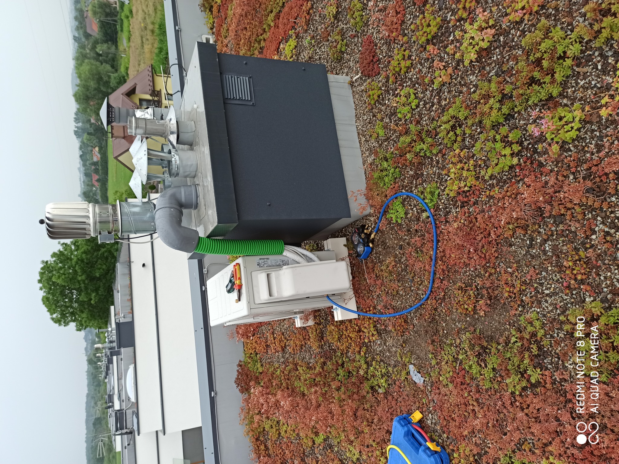 Jednostka zewnętrzna klimatyzatora na dachu pokrytym roślinnością, podłączona do manometrów za pomocą niebieskiego węża, obok leżą narzędzia i niebieska skrzynka narzędziowa.