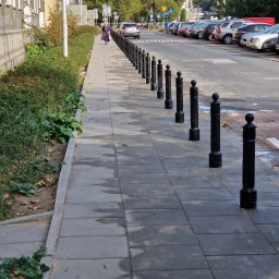 PROBRUK - Szary chodnik z kostki brukowej oddzielony rzędem czarnych słupków od ulicy z zaparkowanymi samochodami, w tle budynek i zieleń.