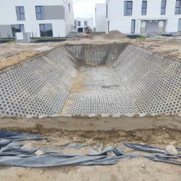 Wykop pod zbiornik retencyjny wyłożony ażurowymi płytami betonowymi w kształcie trójkątów, na tle budynków mieszkalnych w trakcie budowy, z widoczną koparką w oddali.