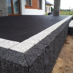 PROBRUK - Nowoczesny taras z czarnej kostki brukowej obramowanej jasnoszarą kostką, z niskim murem oporowym z ciemnej kostki i schodami, na tle domu z ceglaną elewacją.