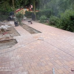 PROBRUK - Trwa układanie chodnika z kostki brukowej w ogrodzie, widoczne przygotowane podłoże, częściowo ułożona nawierzchnia z brązowo-beżowej kostki, pracownik z wózkiem oraz betoniarka w tle.