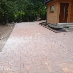 Nowo ułożona, beżowo-brązowa nawierzchnia z kostki brukowej prowadząca do drewnianego domu z widocznymi schodami wejściowymi, z fragmentem terenu przygotowanego pod dalsze prace brukarskie po lewej...