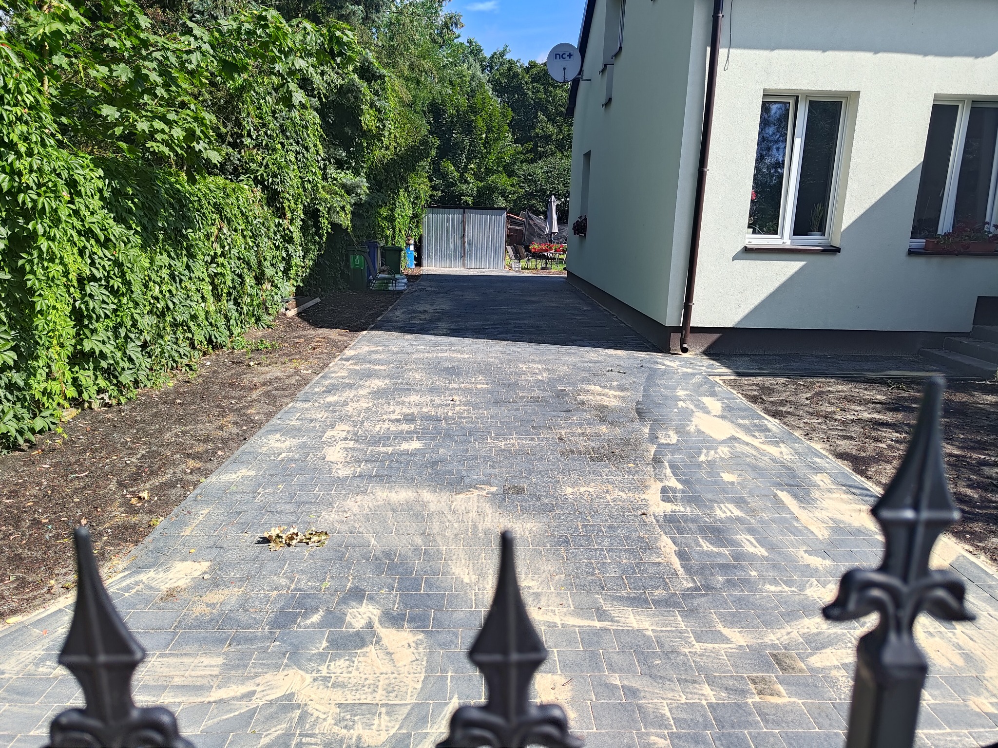 Ułożona szara kostka brukowa prowadząca do garażu blaszanego, widok zza ozdobnego, żeliwnego ogrodzenia, z zielonym żywopłotem po lewej stronie i jasnym budynkiem po prawej.