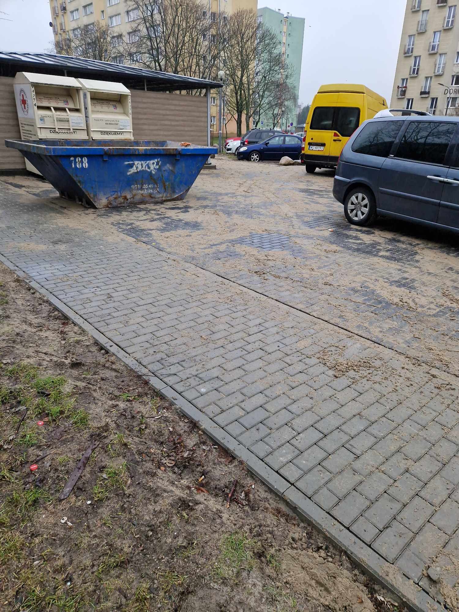Szara kostka brukowa pokryta piaskiem na tle miejskiego krajobrazu z kontenerem na śmieci i zaparkowanymi samochodami.