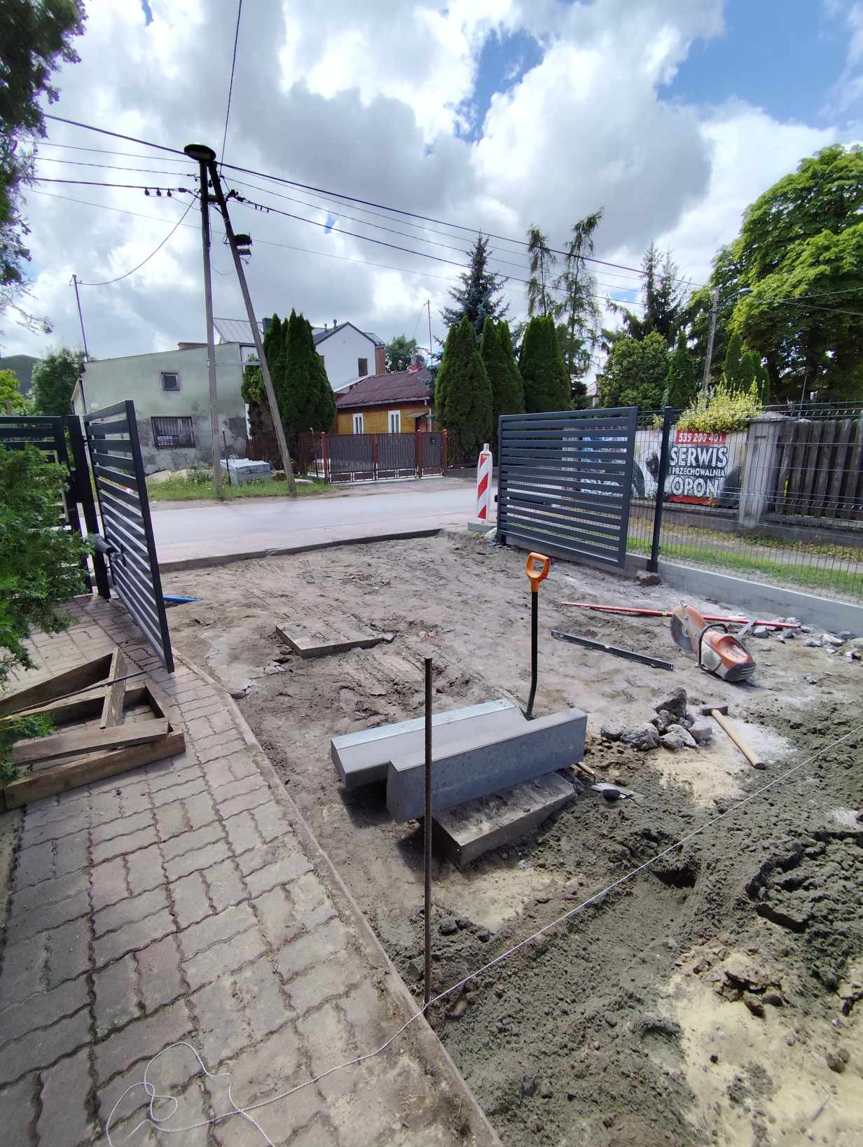 Montaż bramy przesuwnej z poziomymi profilami, widoczne elementy fundamentu pod bramę, wykop, sznurek wyznaczający linię, piła do cięcia betonu i łopata w tle, w tle domy mieszkalne.