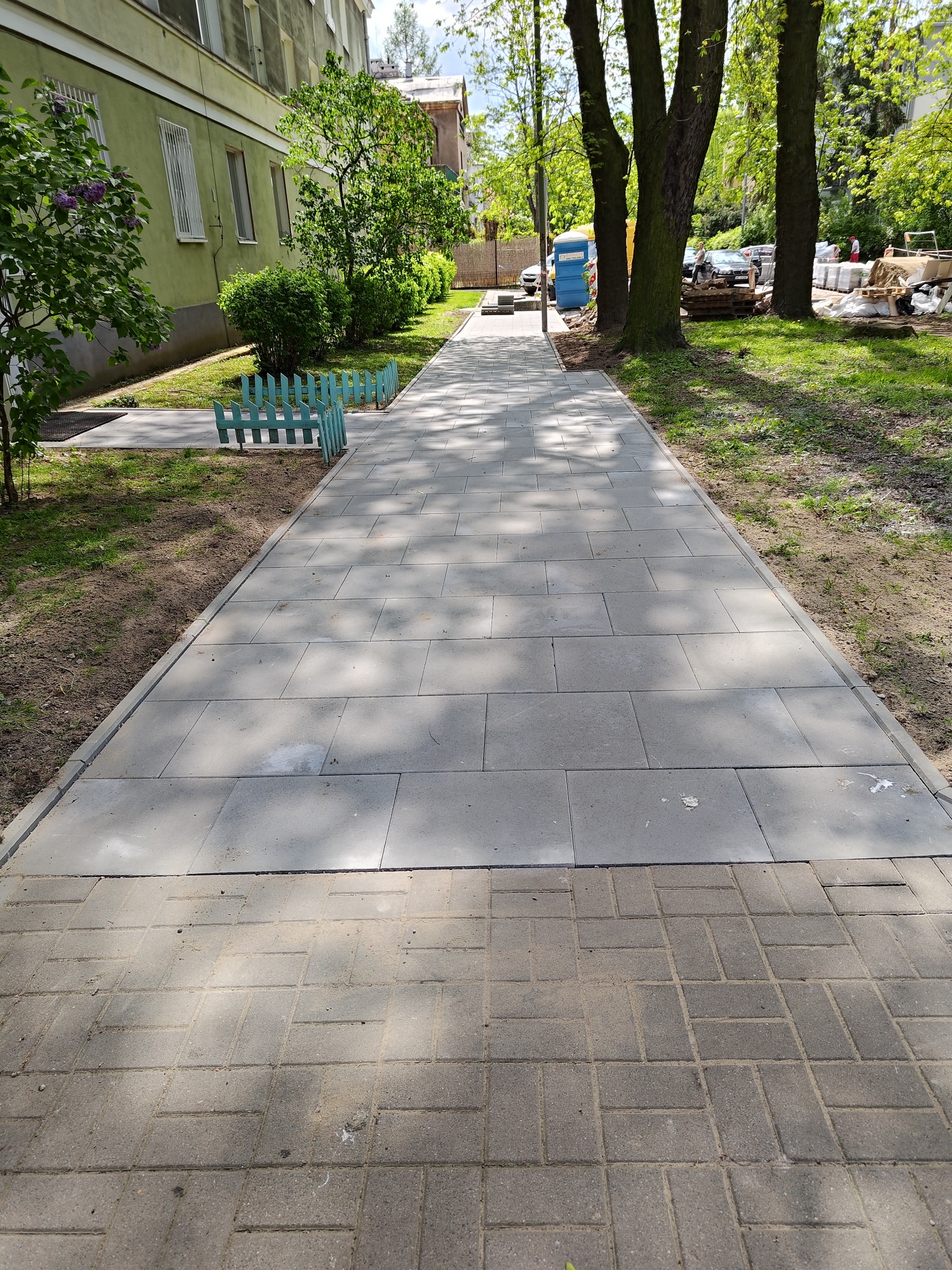 Długa ścieżka z szarych płyt chodnikowych prowadząca wzdłuż budynku z zieloną elewacją, z fragmentem starej nawierzchni z kostki na pierwszym planie i materiałami budowlanymi w tle.