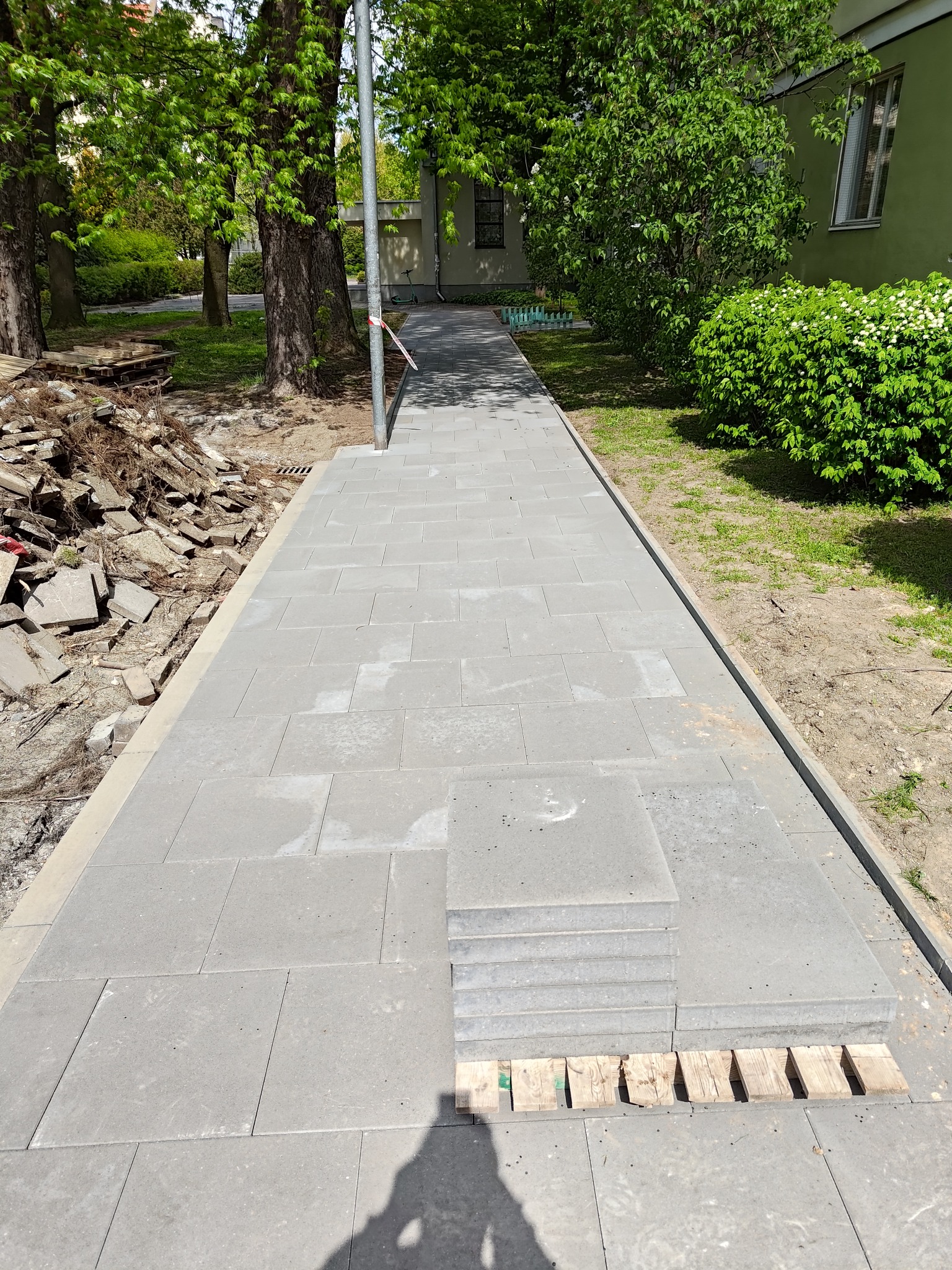 Układanie szarej kostki brukowej na ścieżce, paleta z kostką na pierwszym planie, gruz po prawej stronie, w tle zieleń i budynek.