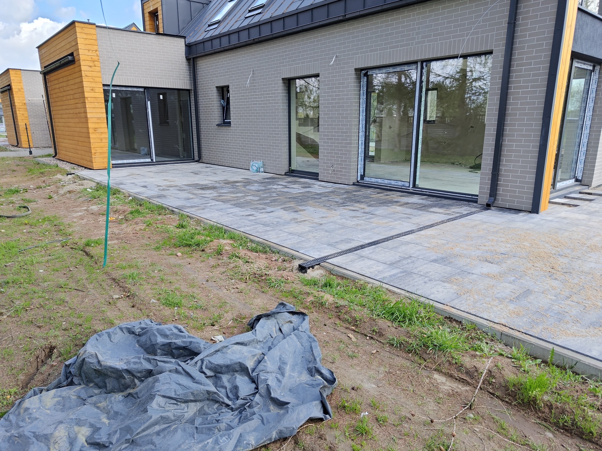 Nowoczesny dom z szarej cegły z dużymi przesuwnymi oknami, wyłożony szarą kostką brukową z odpływem liniowym, widoczna czarna plandeka na ziemi.