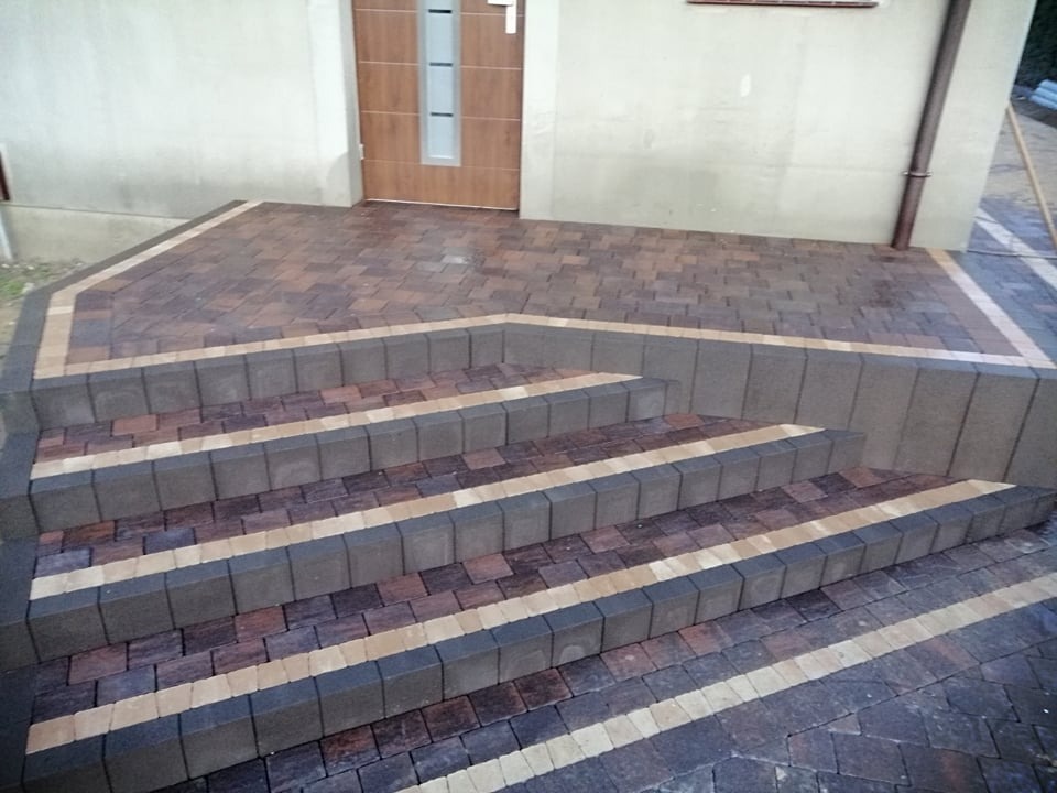 Zewnętrzne schody z kostki brukowej w odcieniach brązu i szarości prowadzące do drzwi wejściowych budynku z elewacją w jasnym kolorze.