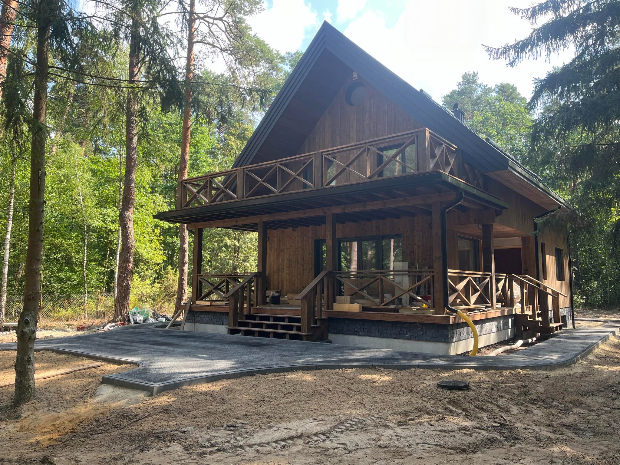 Nowoczesny, drewniany dom z tarasem i balkonem w otoczeniu lasu, z wykończoną nawierzchnią z szarej kostki brukowej.