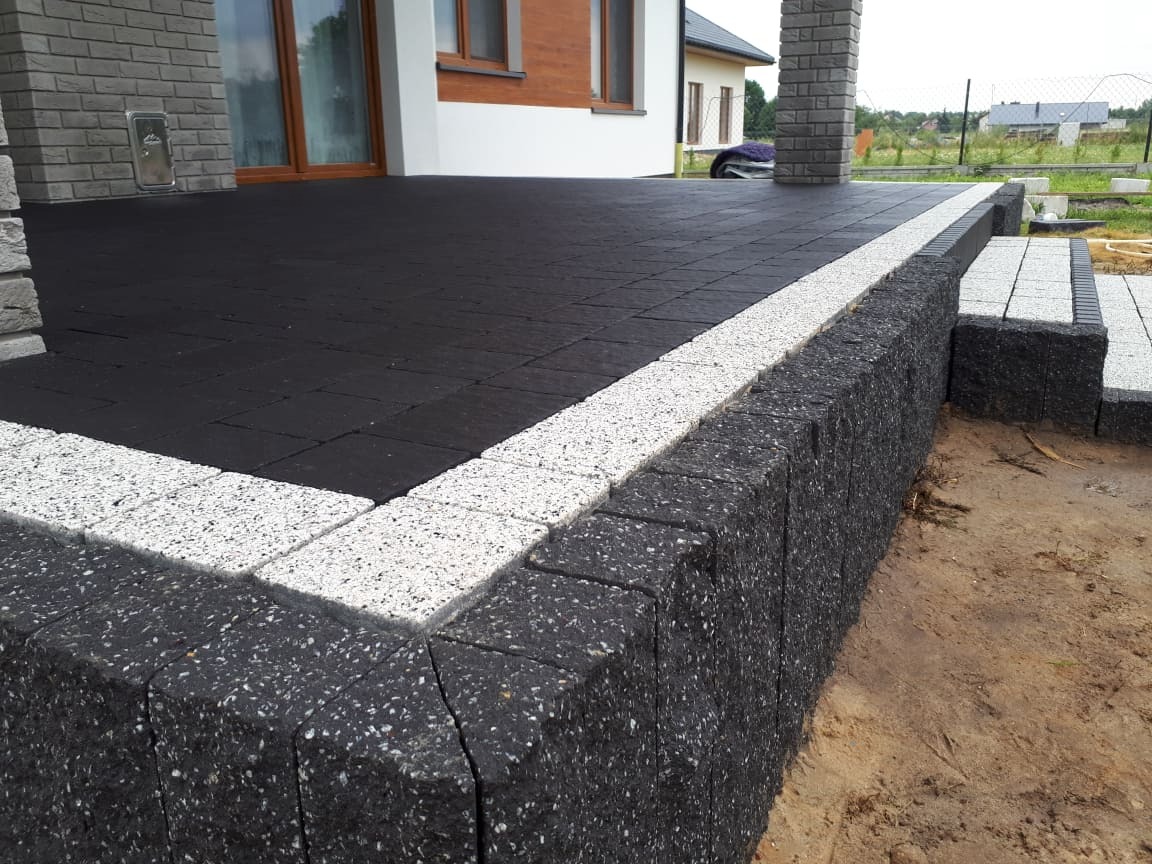 Nowoczesny taras z czarnej kostki brukowej obramowanej jasnoszarą kostką, z niskim murem oporowym z ciemnej kostki i schodami, na tle domu z ceglaną elewacją.