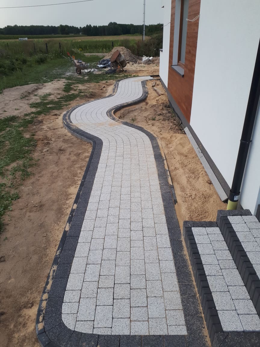 Ścieżka z jasnoszarej kostki brukowej z ciemnoszarą obwódką, wijąca się od schodów domu w kierunku trawnika; w tle widoczna taczka i betoniarka.