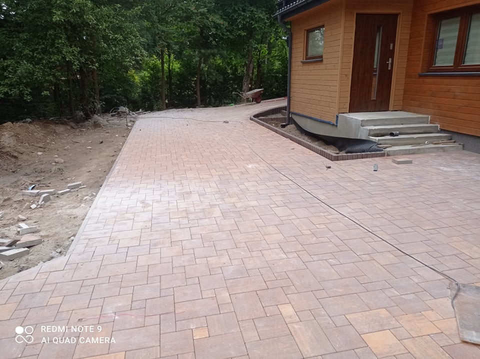 Nowo ułożona, beżowo-brązowa nawierzchnia z kostki brukowej prowadząca do drewnianego domu z widocznymi schodami wejściowymi, z fragmentem terenu przygotowanego pod dalsze prace brukarskie po lewej...
