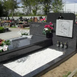 Polerowany, czarny nagrobek z jasnymi kamykami wewnątrz, ozdobiony rzeźbą Matki Boskiej, kwiatami i zniczami na tle cmentarza.