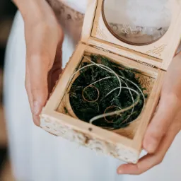 Dłonie trzymają otwarte, drewniane pudełko z wygrawerowanymi inicjałami i datą, w kt&oacute;rym na zielonym mchu leżą dwie złote obrączki.