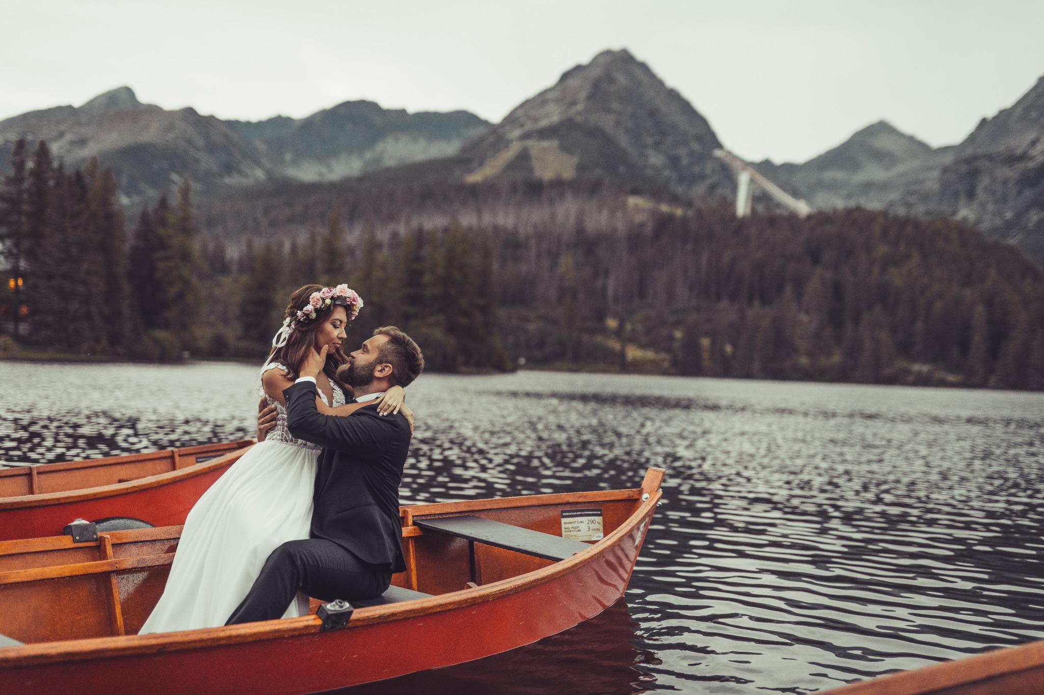 Para młoda w łódce na jeziorze z górami w tle, panna młoda ma wianek z różowych kwiatów, pan młody w ciemnym garniturze.