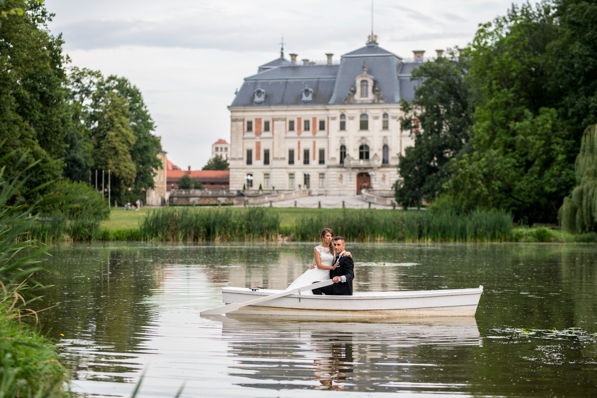 Para młoda w łódce na jeziorze, w tle pałac w stylu barokowym.
