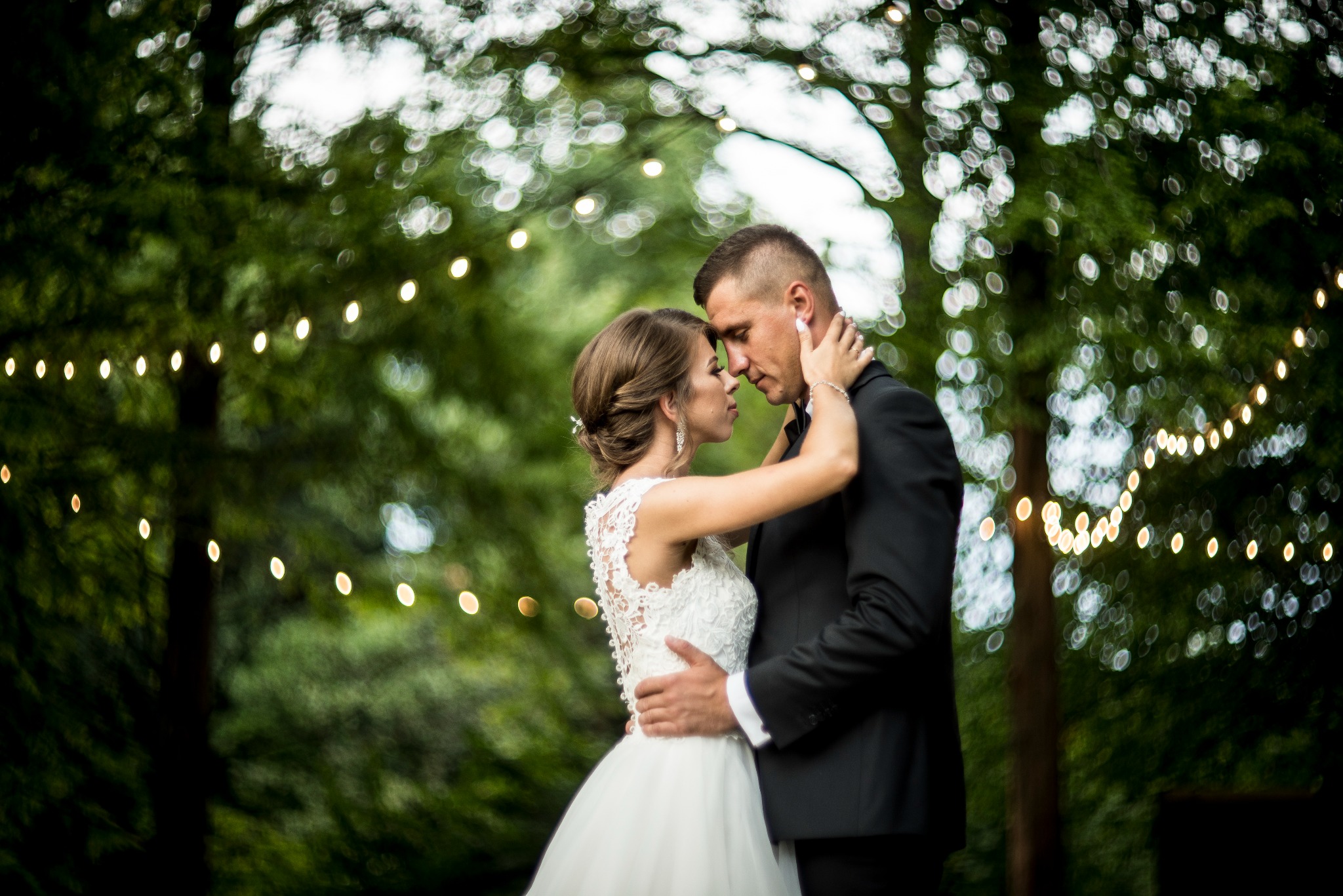 Para młoda w objęciach, panna młoda w koronkowej sukni, pan młody w czarnym garniturze, na tle drzew i dekoracyjnych światełek.