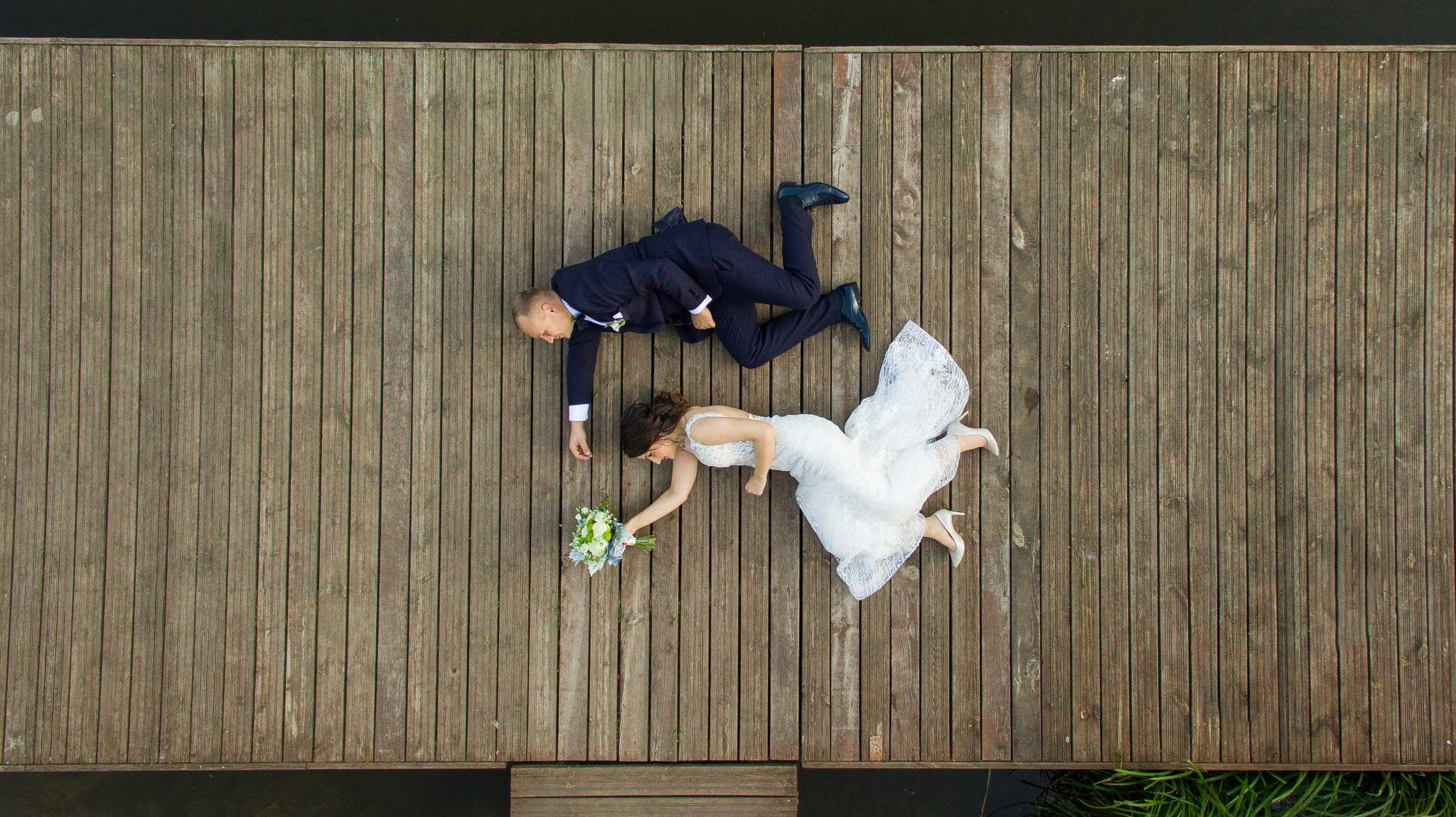 Para młoda leżąca na drewnianym pomoście nad wodą, widok z góry: panna młoda w białej sukni z bukietem i pan młody w granatowym garniturze.