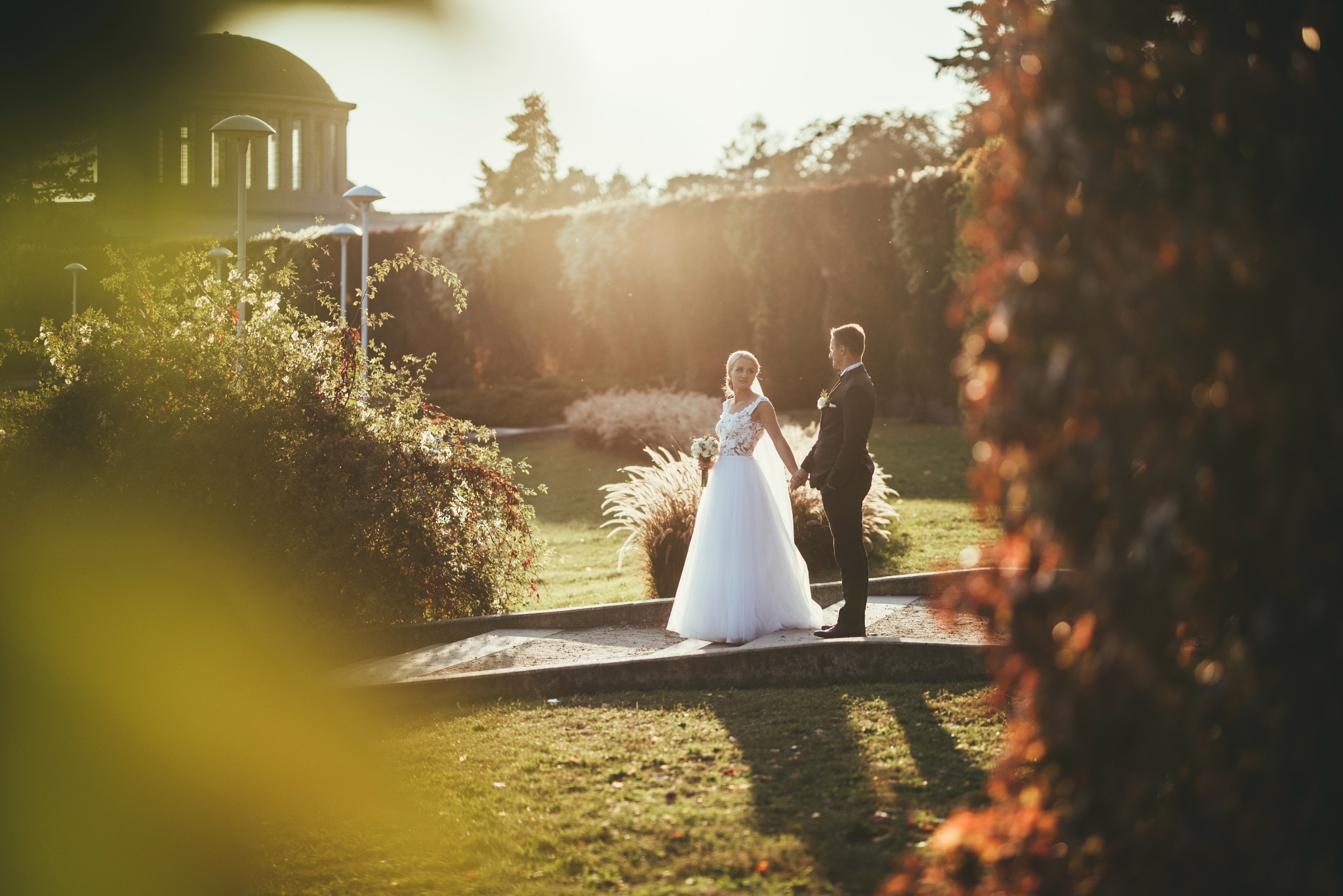 Uśmiechnięta panna młoda w koronkowej sukni i welonie, trzymająca bukiet, spaceruje z panem młodym w ciemnym garniturze po parku w promieniach zachodzącego słońca, w tle widoczna architektura parkowa.