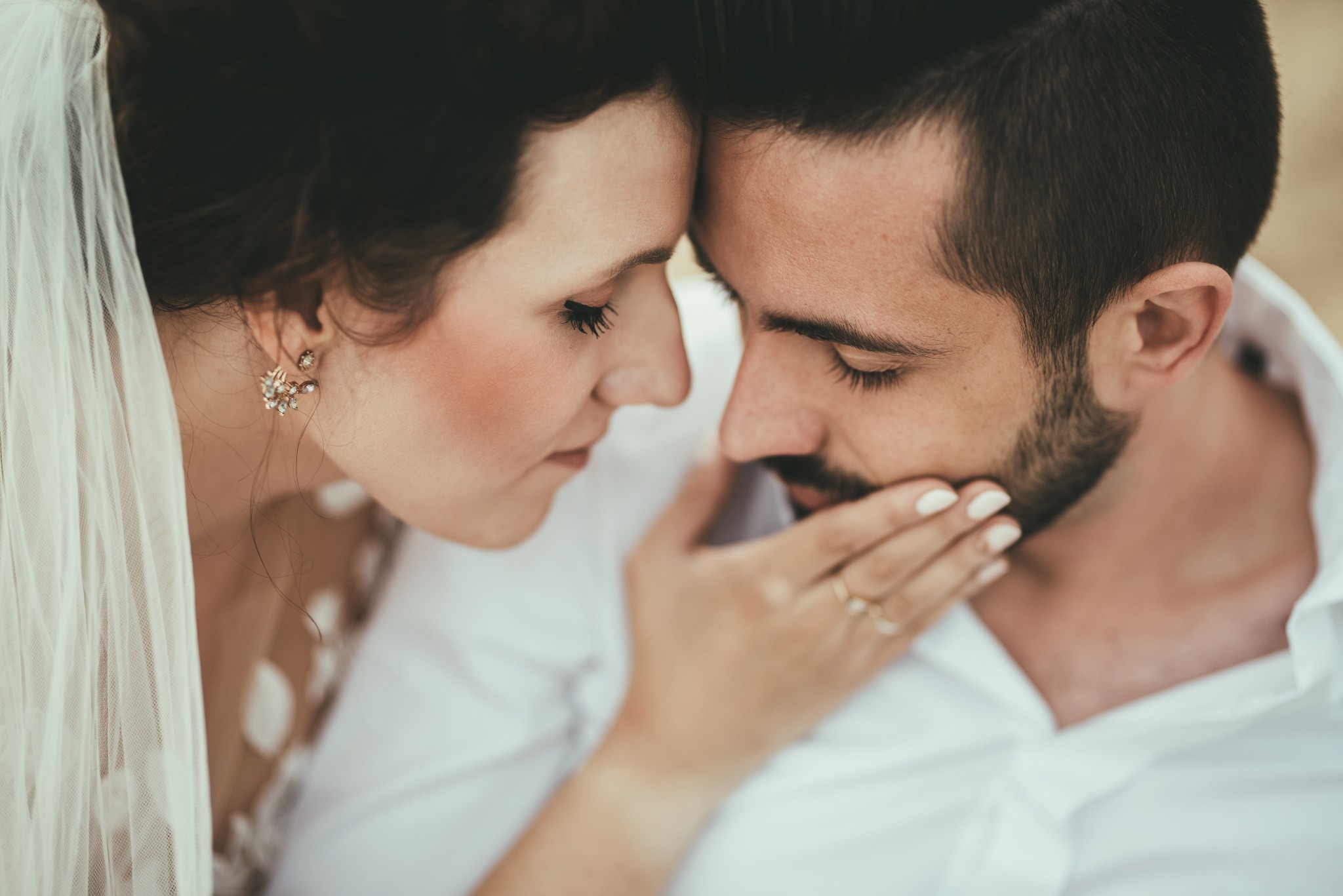 Młoda para, zbliżone ujęcie, panna młoda w welonie i z delikatną biżuterią, pan młody w białej koszuli, dłoń panny młodej na twarzy pana młodego, intymna chwila.