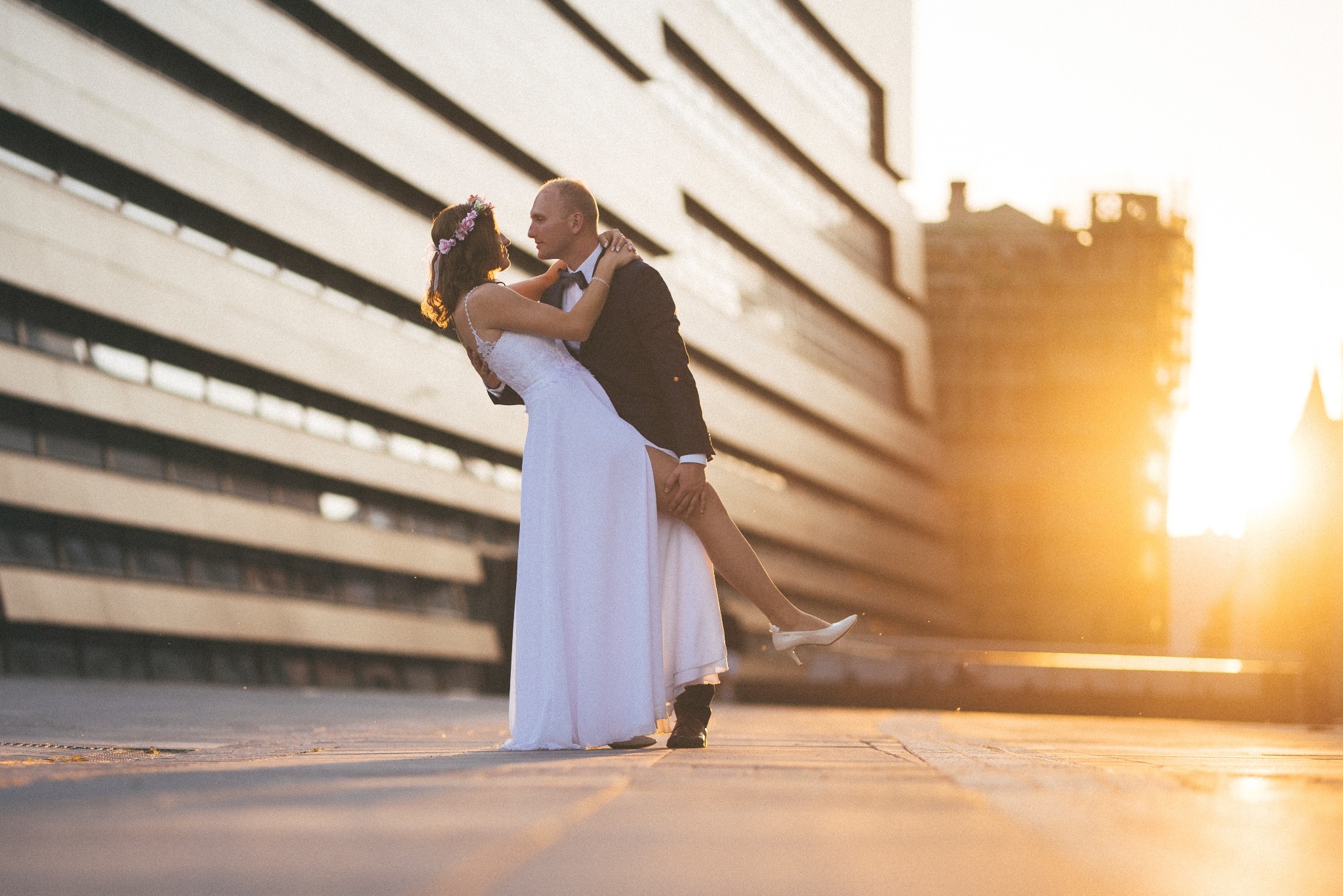 Para młoda w tańcu na tle nowoczesnej architektury w promieniach zachodzącego słońca, panna młoda w białej sukni i wianku, pan młody w ciemnym garniturze i muszce.