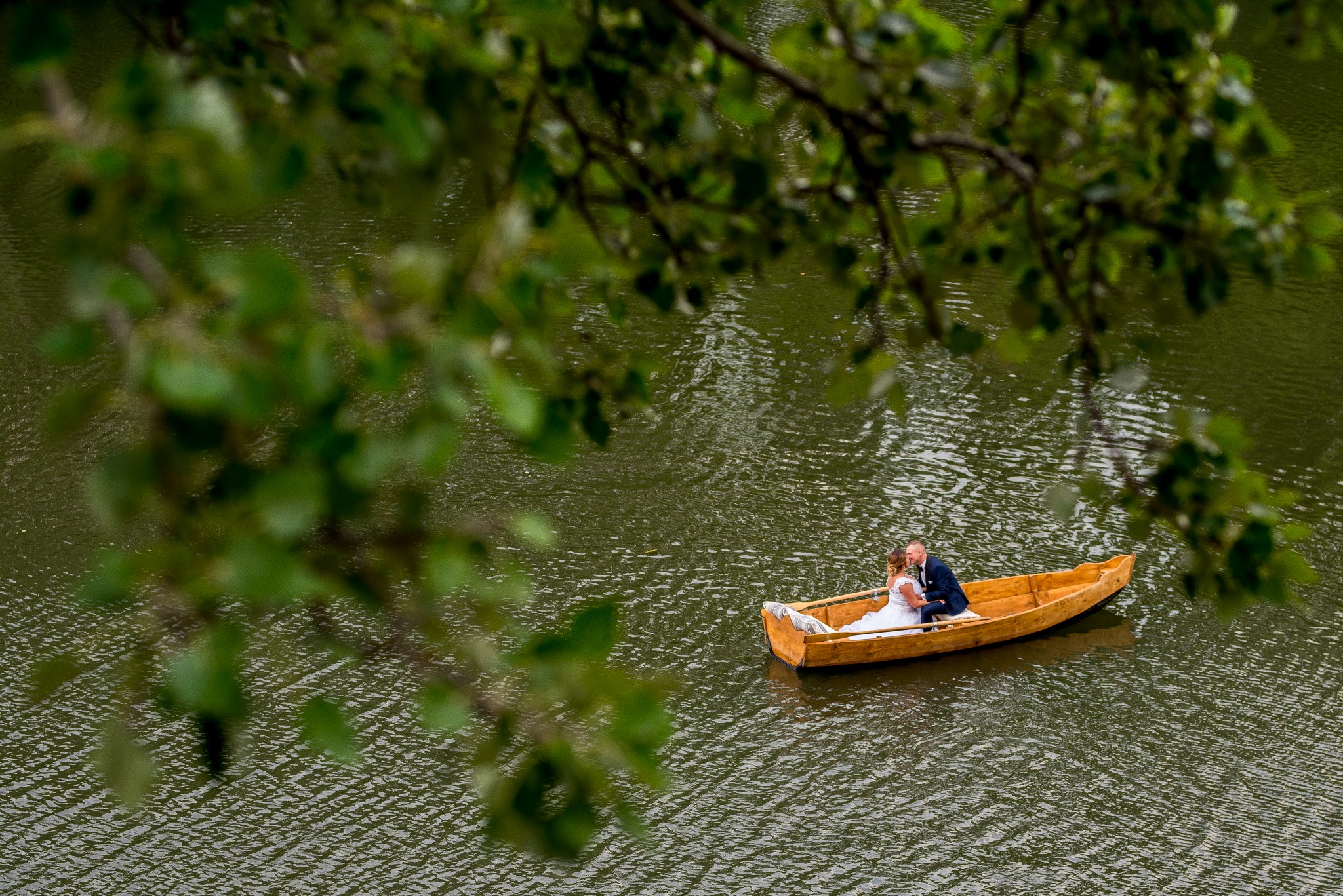 Para młoda w drewnianej łódce na jeziorze, widok z góry przez gałęzie drzew, mężczyzna całuje kobietę w welonie.