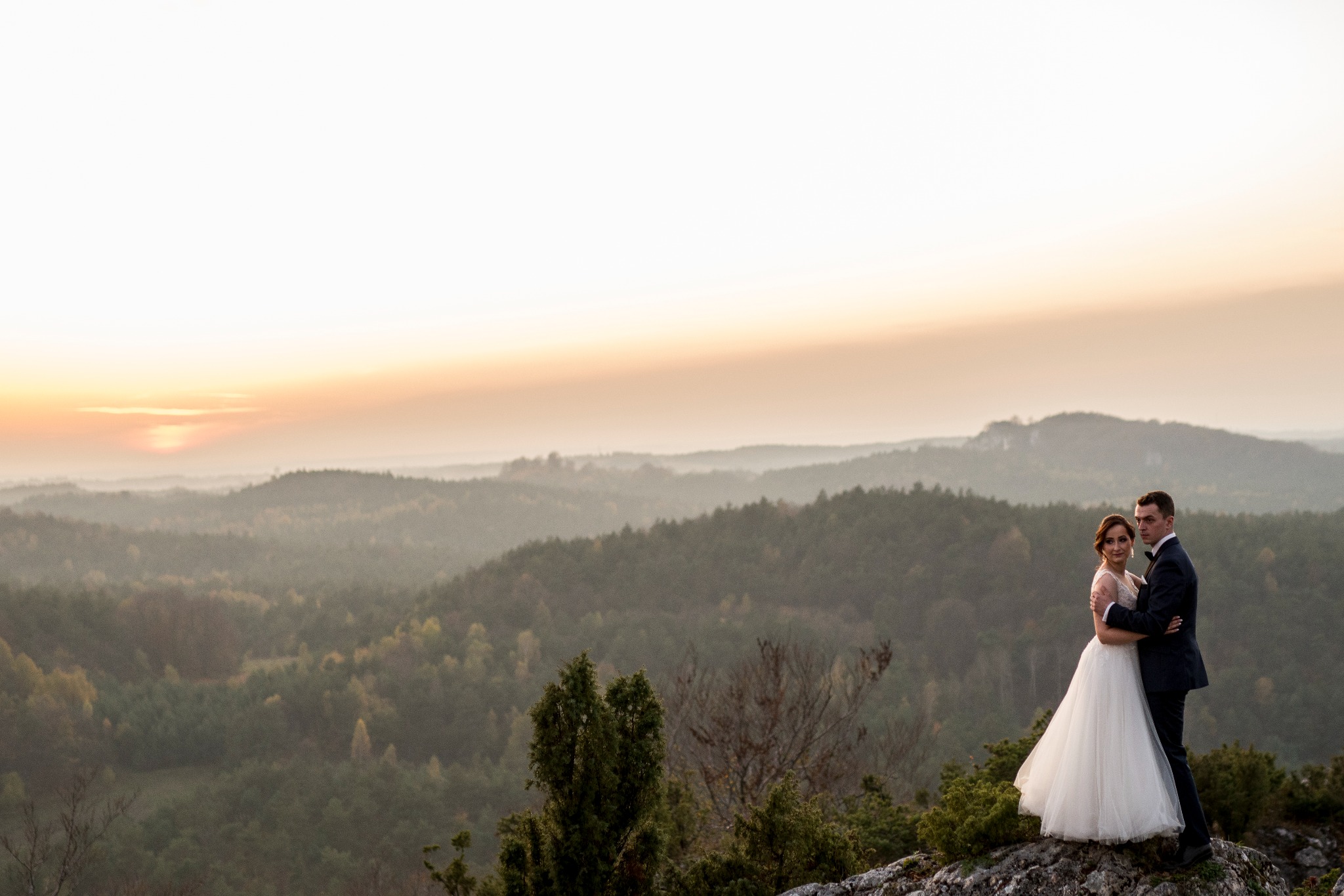 Para młoda w objęciach na skalistym wzgórzu o zachodzie słońca, rozległy widok na zalesione wzgórza w oddali.