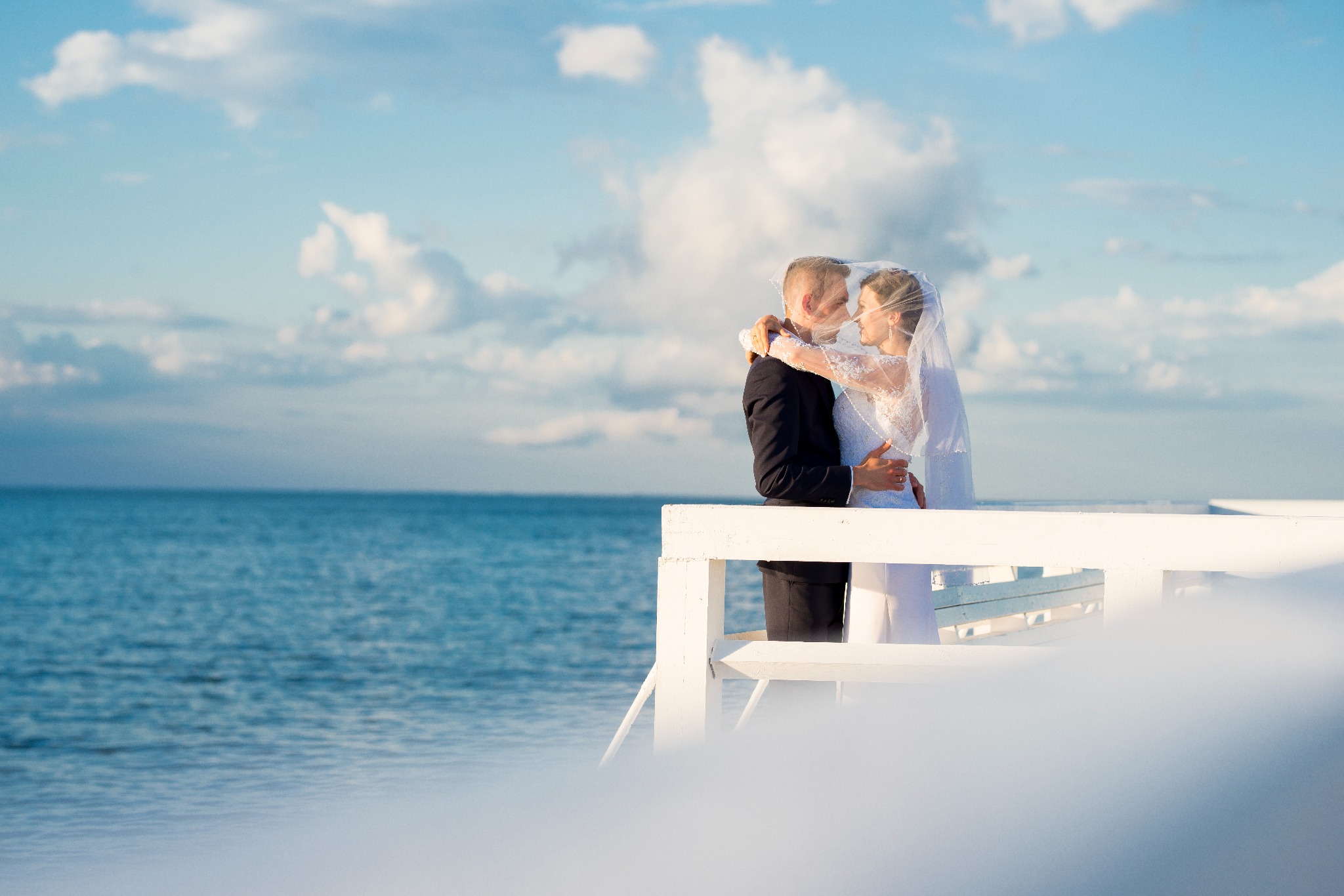 Para młoda, objęta welonem, stoi na białym pomoście z widokiem na morze i błękitne niebo z chmurami.