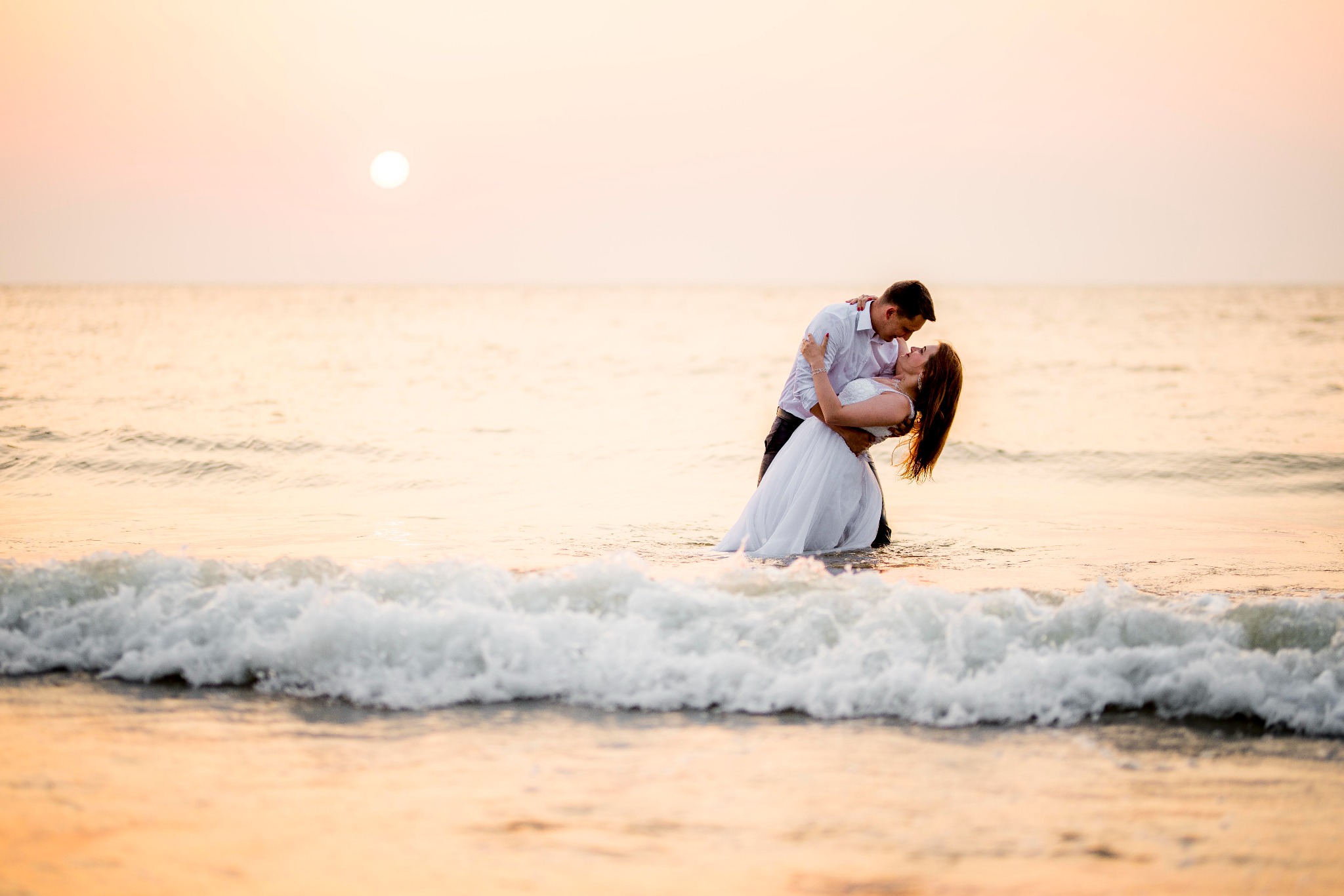 Para młoda w objęciach stoi w morzu podczas zachodu słońca, fale delikatnie obmywają ich stopy.