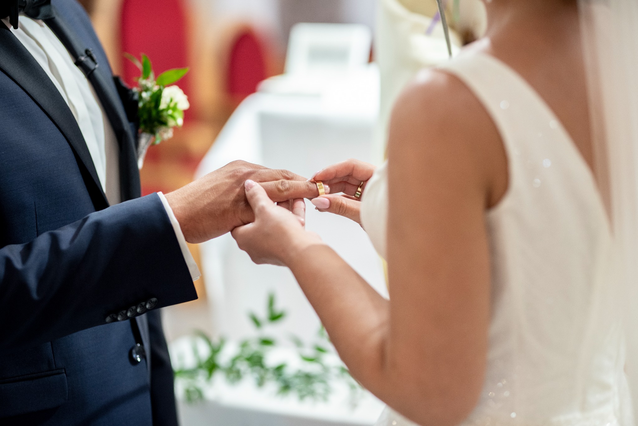 Panna Młoda zakłada złotą obrączkę na palec Pana Młodego podczas ceremonii ślubnej, w tle butonierka z białych kwiatów i zielonych liści w klapie granatowego garnituru.