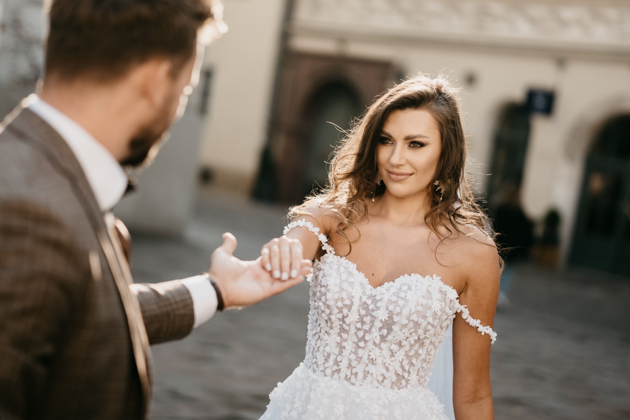 Panna młoda w białej sukni z aplikacjami kwiatowymi, trzymająca za rękę pana młodego w brązowym garniturze, na tle miejskiej architektury w ciepłym świetle słonecznym.