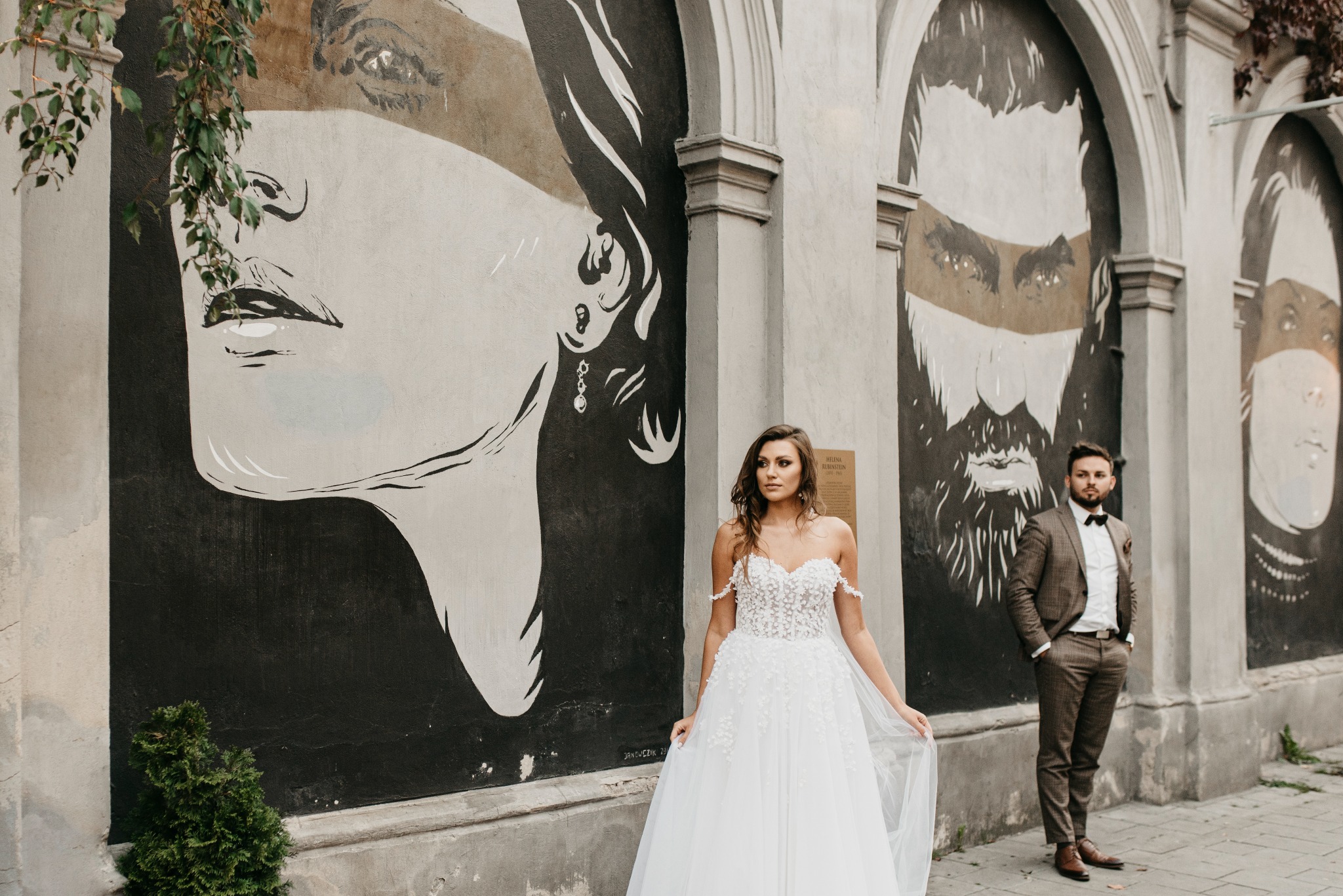 Panna młoda w koronkowej sukni ślubnej pozuje z panem młodym w brązowym garniturze na tle ściany z muralami portretów