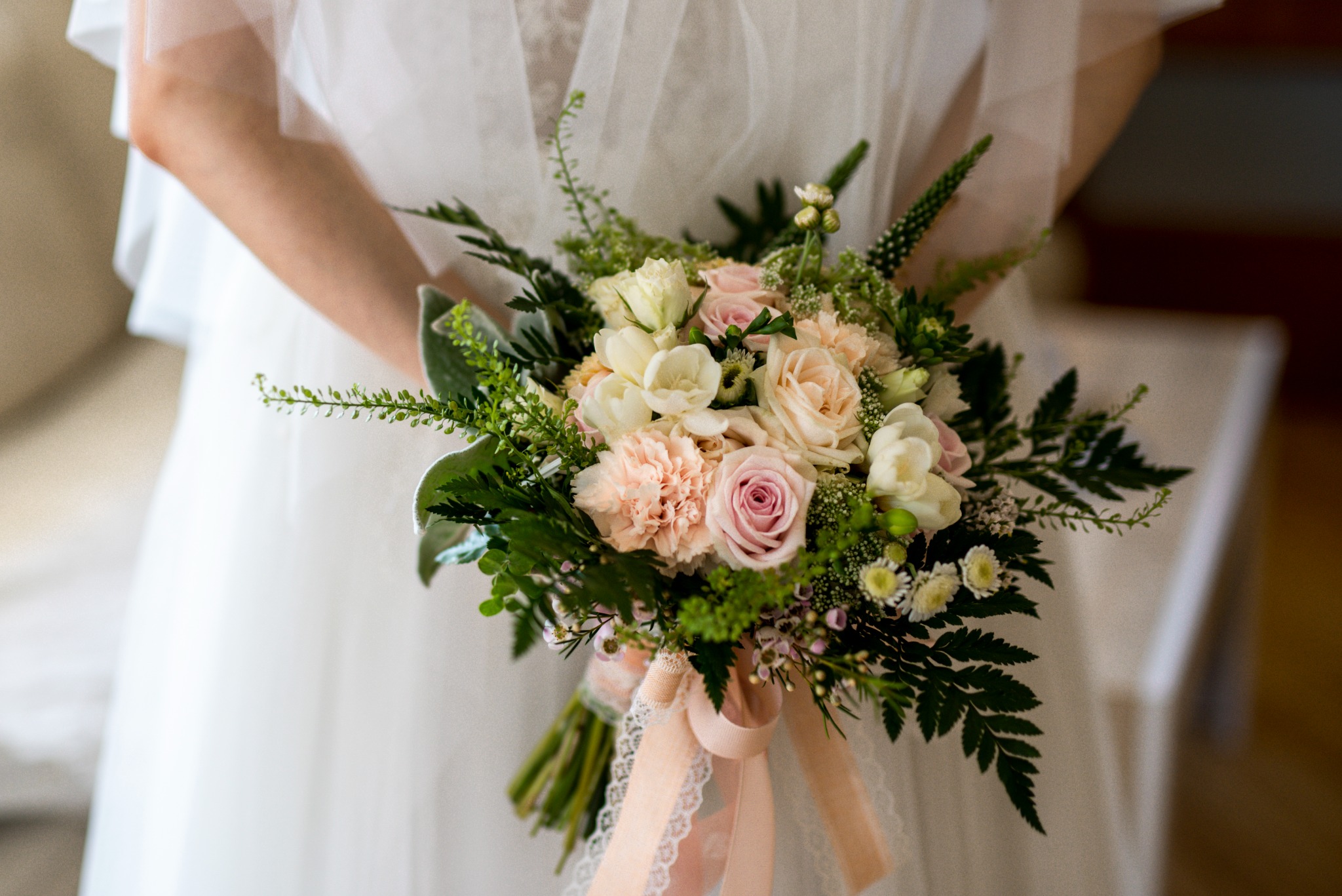 Panna młoda trzyma bukiet ślubny z różowych i białych róż oraz zielonych dodatków, przewiązany kremową wstążką z koronką.