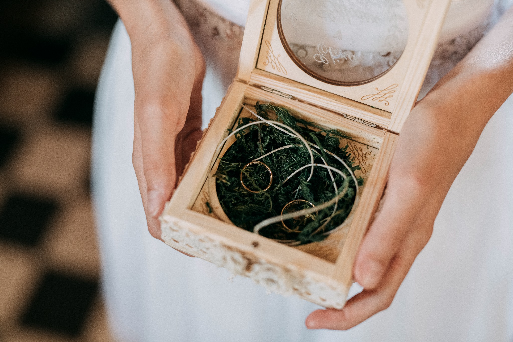 Dłonie trzymają otwarte, drewniane pudełko z wygrawerowanymi inicjałami i datą, w którym na zielonym mchu leżą dwie złote obrączki.