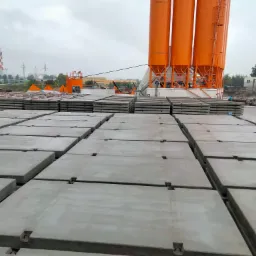 Magazyn betoniarni z ułożonymi płytami drogowymi; w tle pomarańczowe silosy i komin przemysłowy.