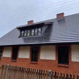 Dwukondygnacyjny dom z czerwonej cegły z nowym, ciemnoszarym dachem z dachówki ceramicznej. Widoczne kominy i okna z ciemnymi ramami. Fragment drewnianego płotu w tle.