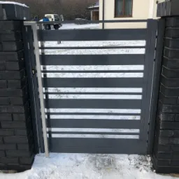 Nowoczesna, szara furtka z poziomymi szczeblami, zamontowana między dwoma słupkami z ciemnej cegły klinkierowej, na tle ośnieżonego podjazdu i budynku mieszkalnego.