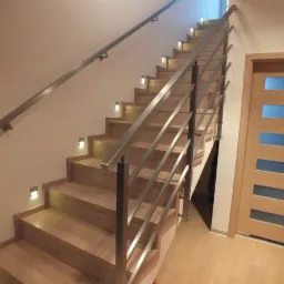 Drewniane schody z podświetleniem stopni i prostą, metalową balustradą, widziane z dołu, z jasną drewnianą podłogą i drzwiami w tle.