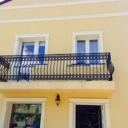 Elewacja budynku z balkonem zabezpieczonym czarną, metalową balustradą o zdobnym wzorze, widok z dołu na tle jasnej, kremowej ściany.