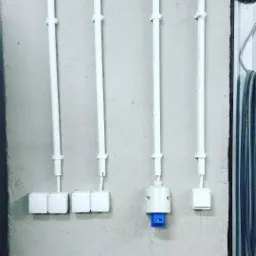 Białe rury instalacyjne przymocowane do szarej ściany, prowadzące do gniazdek i łączników elektrycznych, w tym jedno gniazdko z niebieskim wkładem.