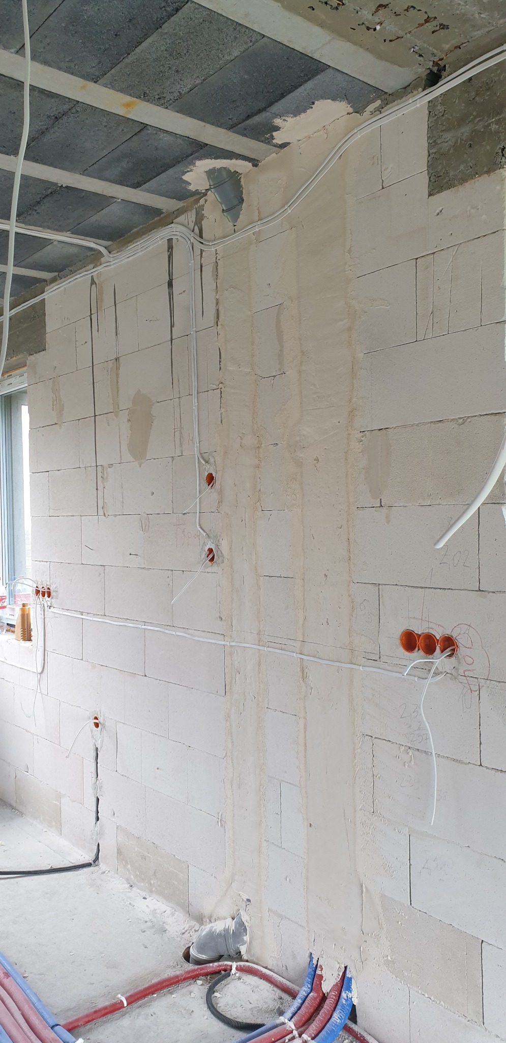 Surowa ściana z betonu komórkowego w trakcie prac instalacyjnych, widoczne kable elektryczne prowadzone natynkowo i w peszlach, puszki instalacyjne w ścianie, oraz rury instalacji wodnej...